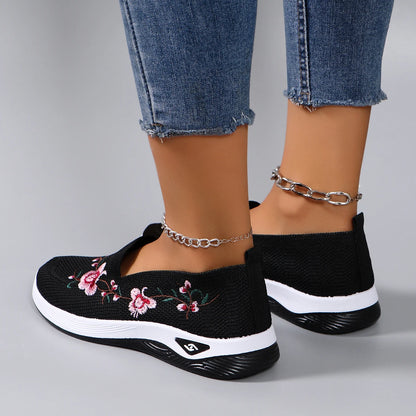 Black slip-on sneakers with floral design worn with blue jeans on a gray background