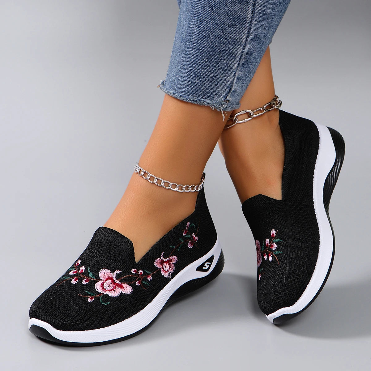Black slip-on sneakers with floral embroidery worn with blue jeans on a gray background