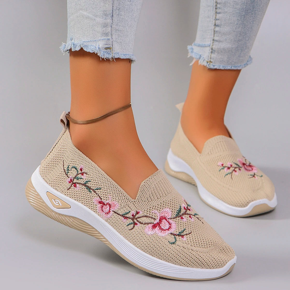 Beige slip-on shoes with floral embroidery worn with light blue jeans on a gray background