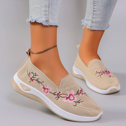 Beige slip-on shoes with floral embroidery worn with light blue jeans on a gray background