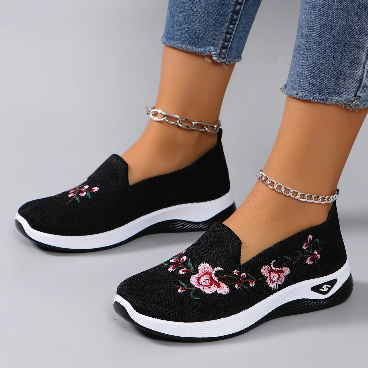 Black slip-on shoes with floral embroidery worn with blue jeans on a gray background