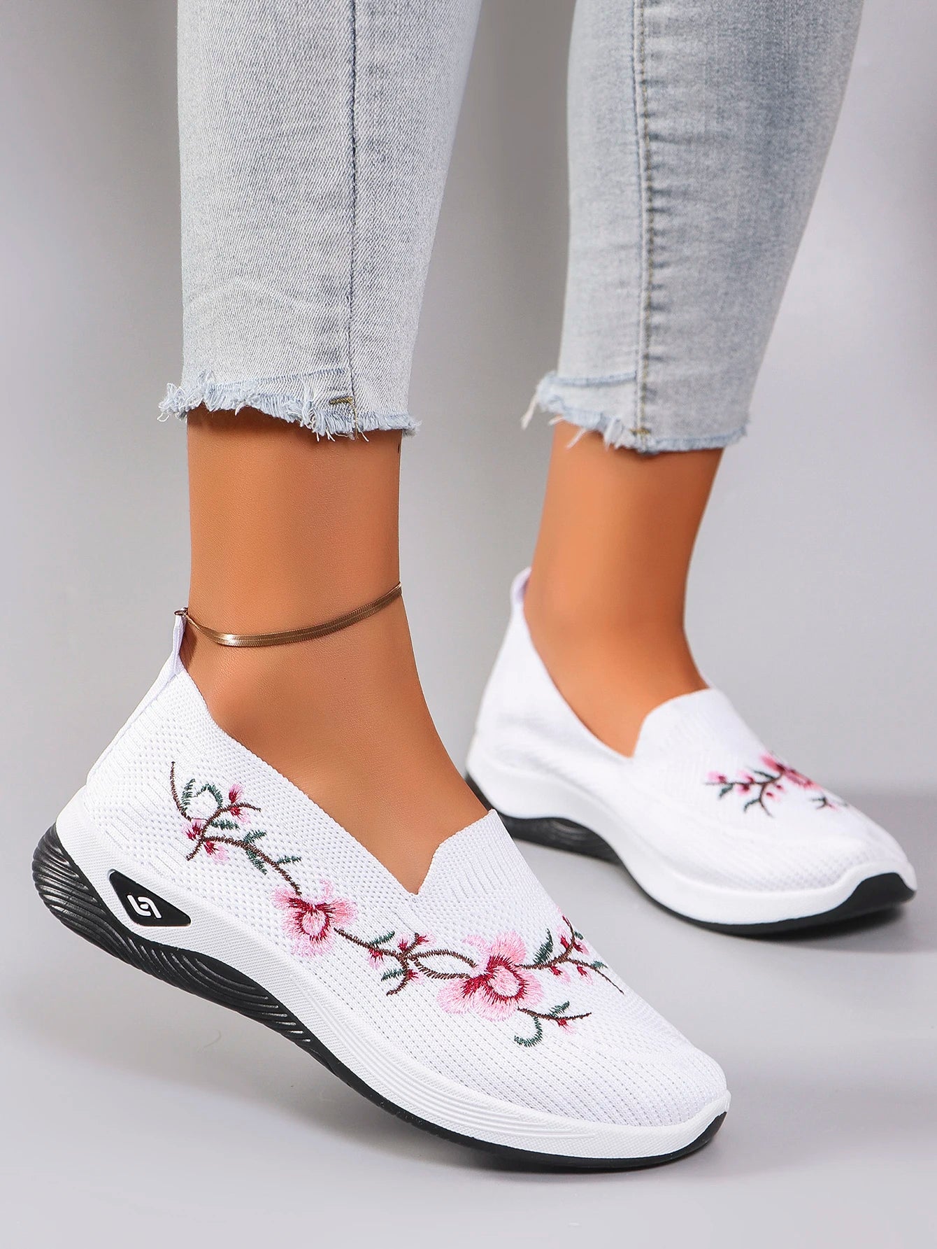 White slip-on shoes with floral embroidery worn with light gray jeans on a neutral background